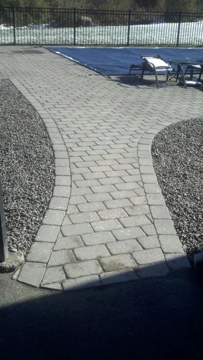 Curved brick paver walkway with herringbone pattern bordered by gravel beds and metal fence