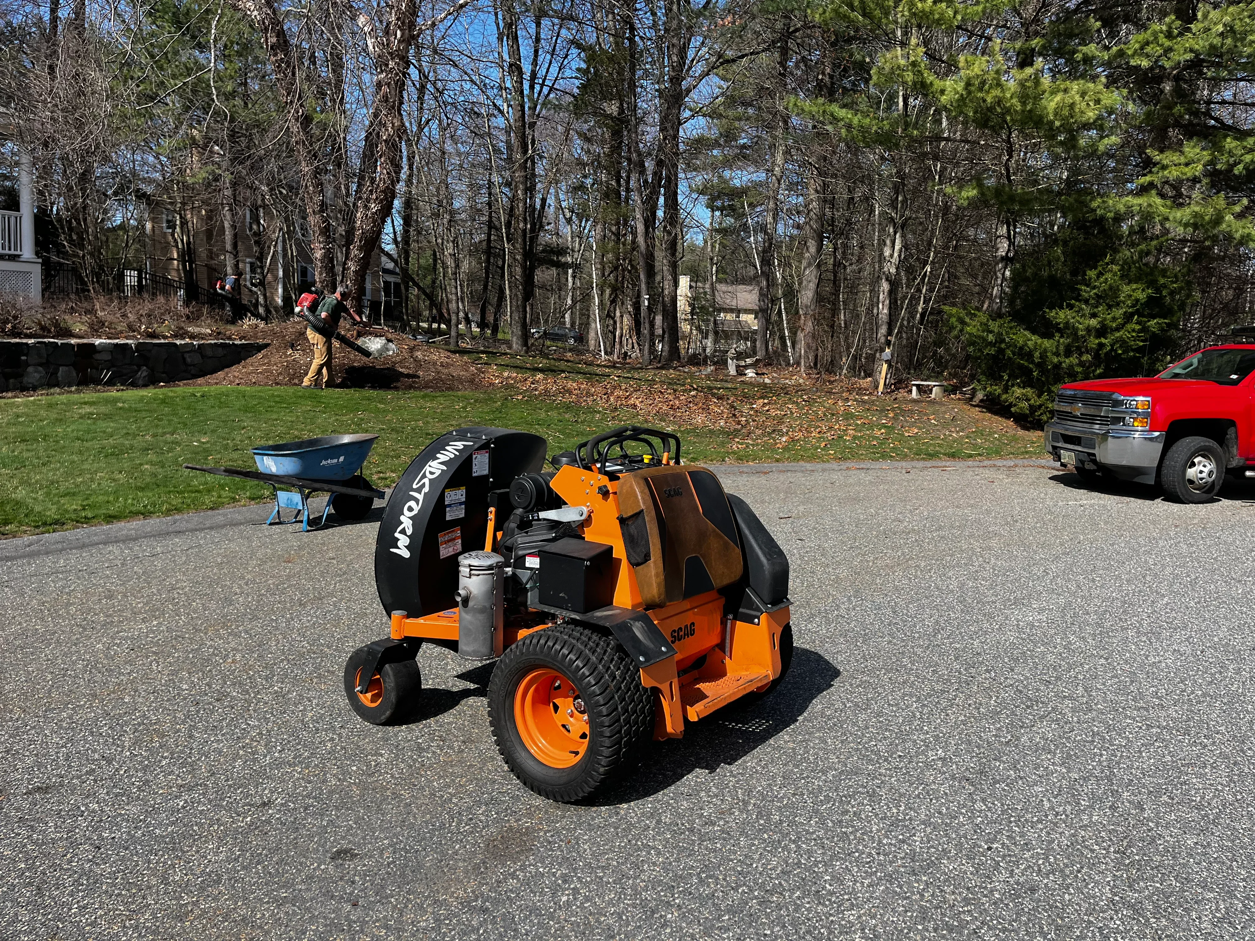 Orange Scag zero-turn mower with attached chipper on residential driveway, crew clearing wooded property in background