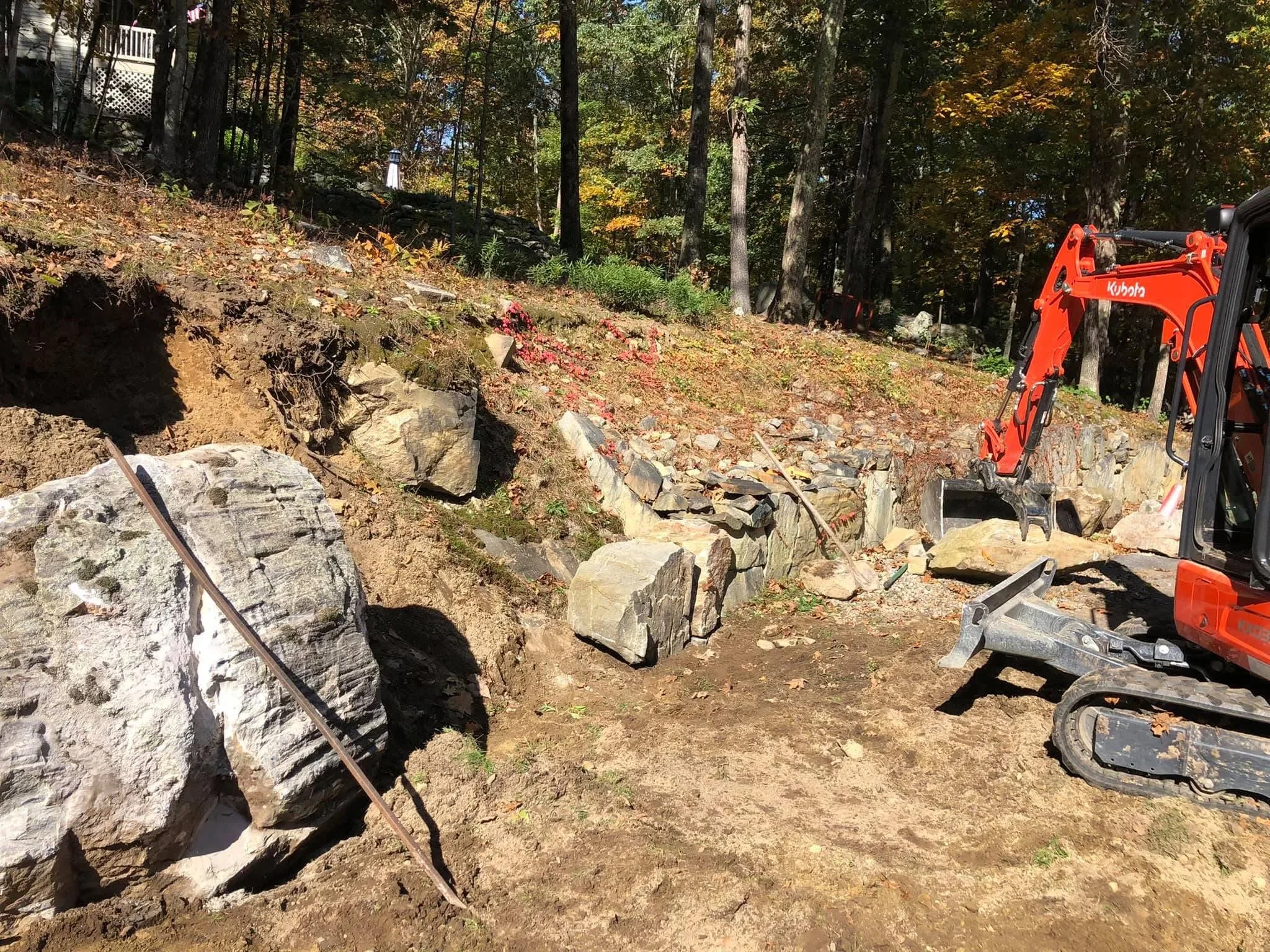 Mini excavator on wooded slope with exposed stone, graded earth, and natural boulders