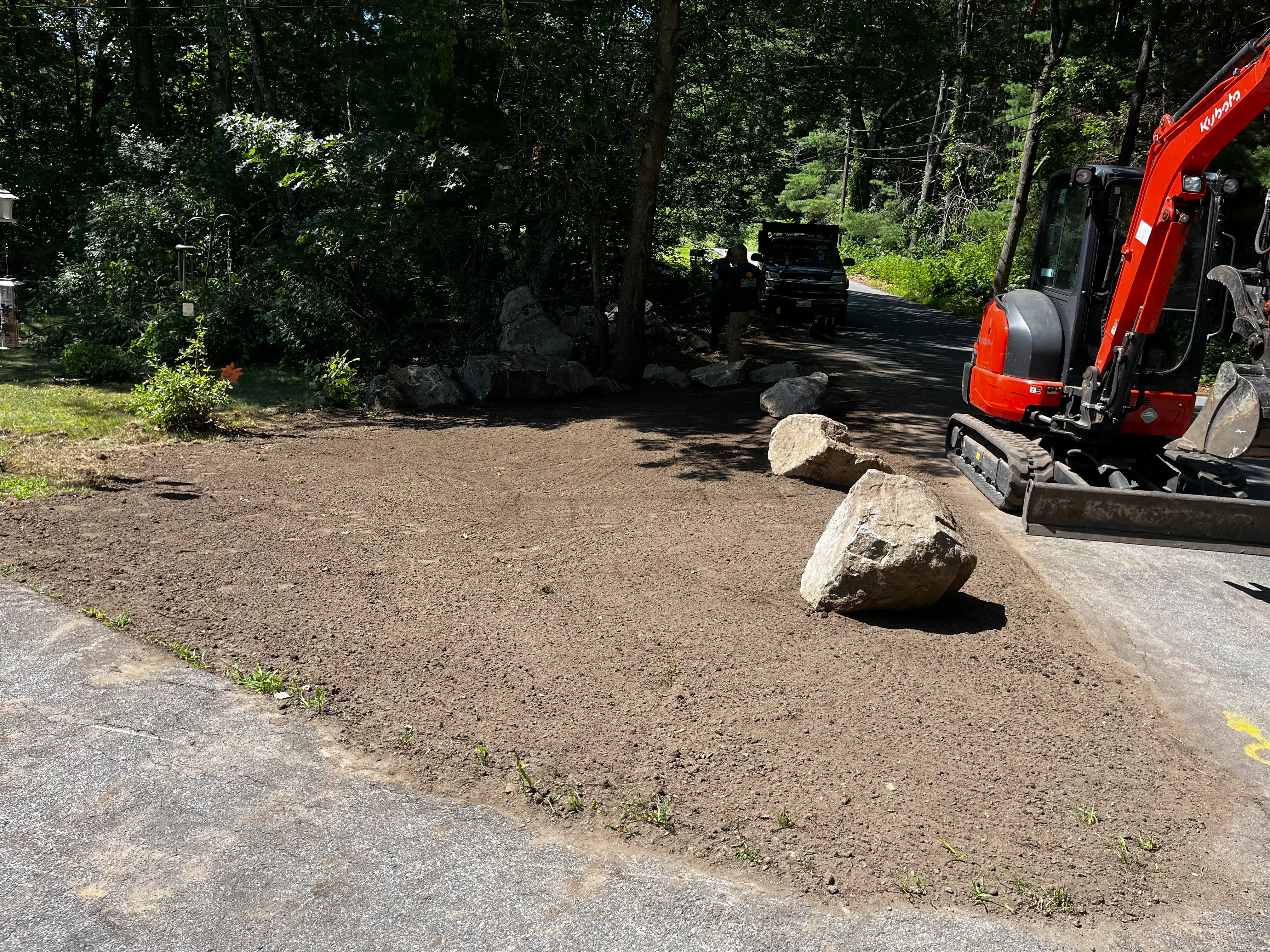 Cleared driveway entrance with positioned boulders and heavy equipment in wooded setting