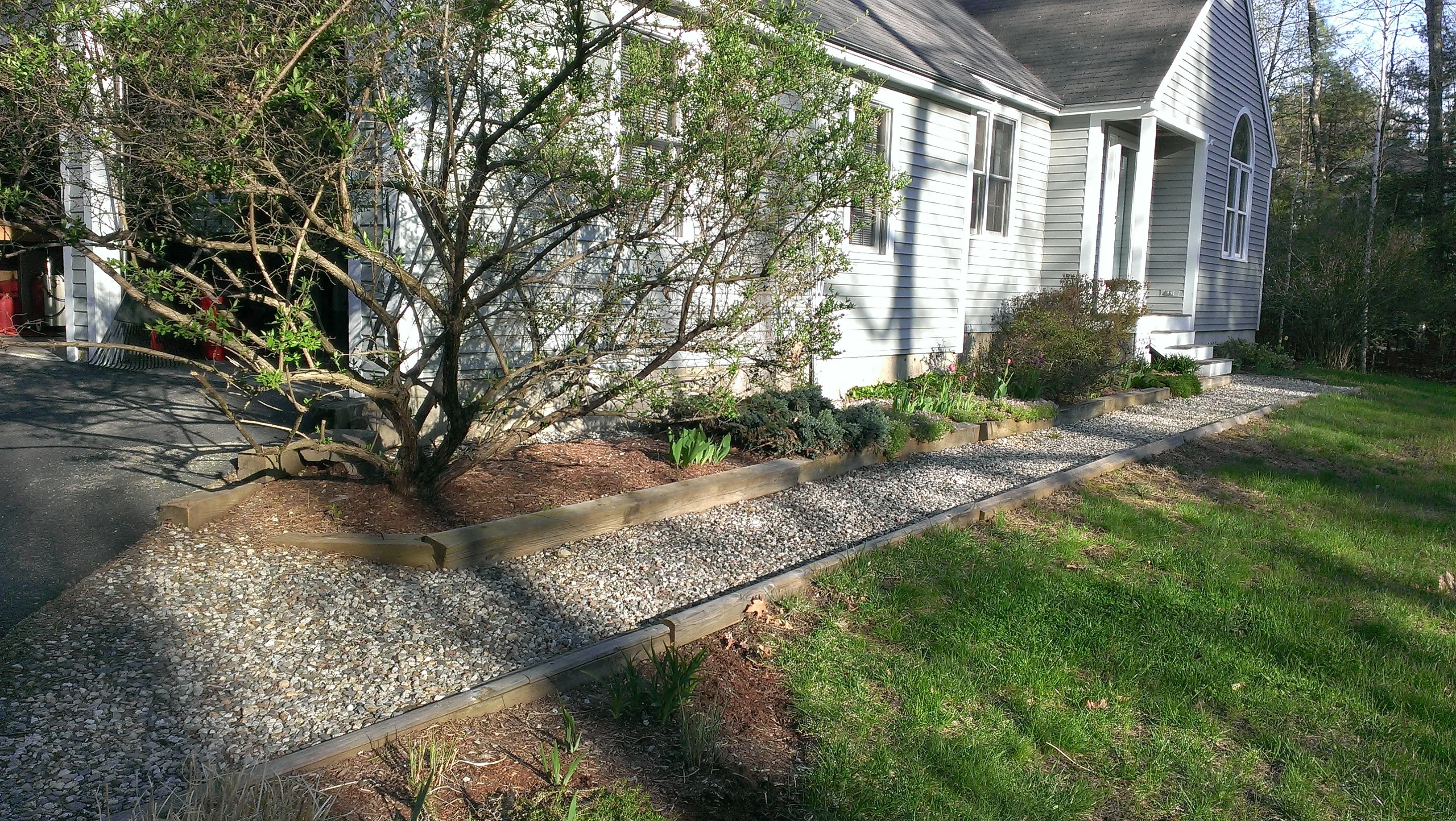 Gravel pathway with timber edging, landscape beds, and mature trees near house foundation