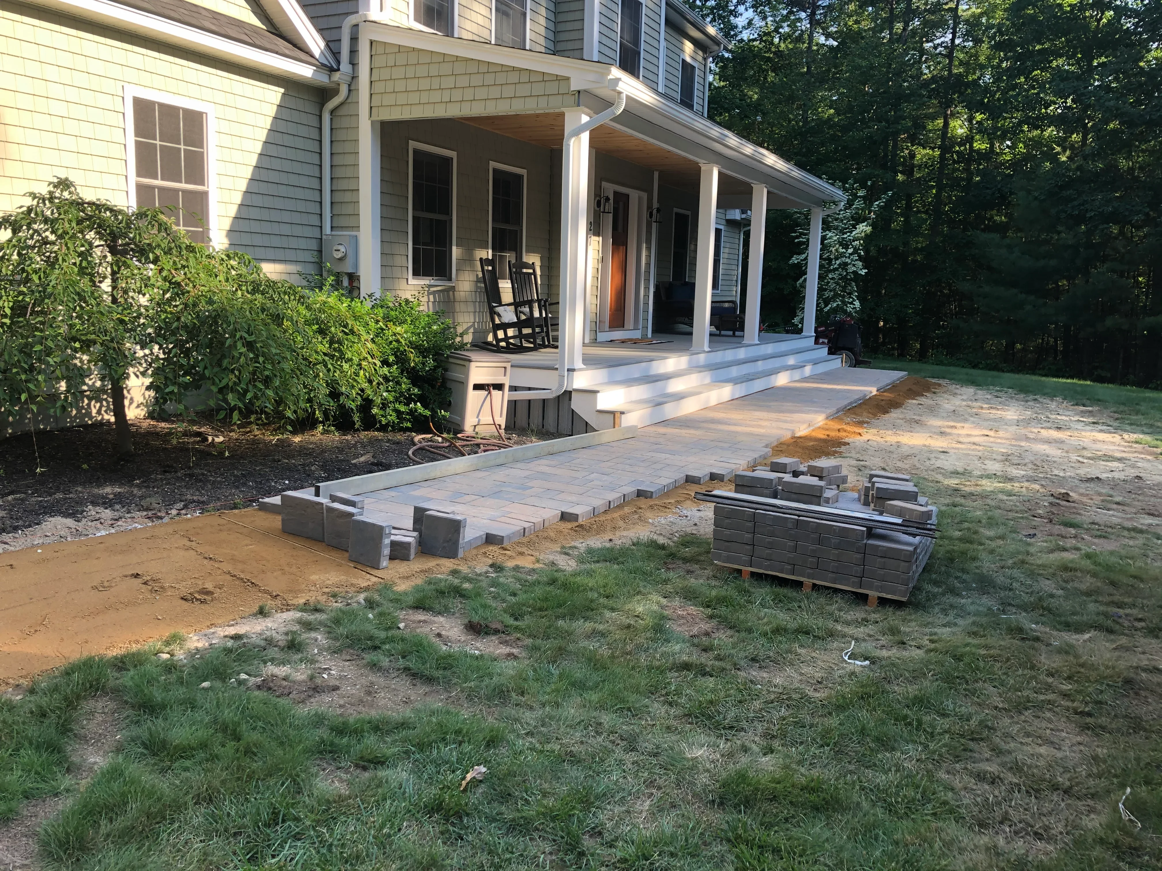 Paver patio installation in progress with concrete base and stacked pavers staged beside home