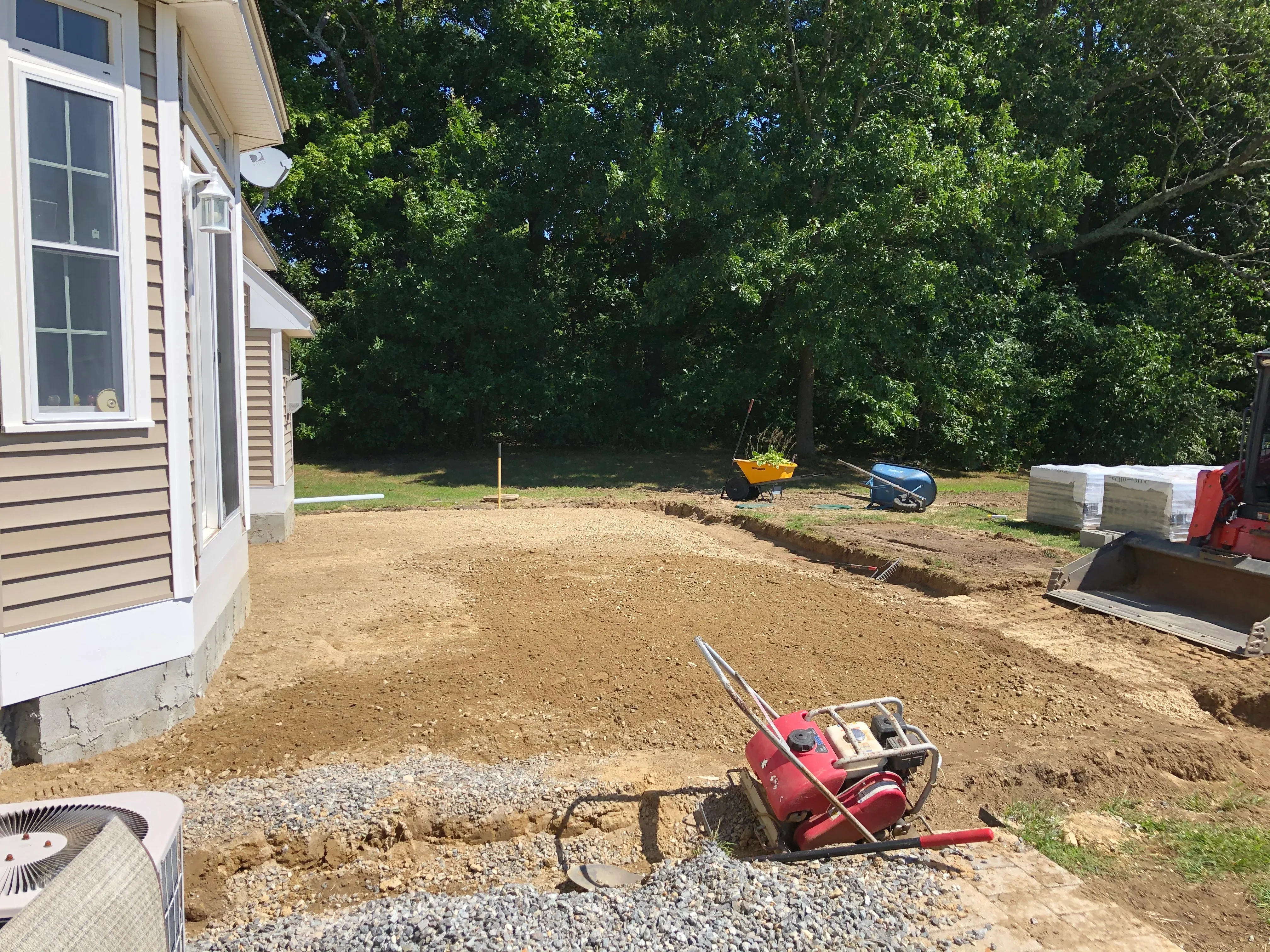 Graded dirt lot with drainage trench, compacted soil base, and construction equipment near residential home
