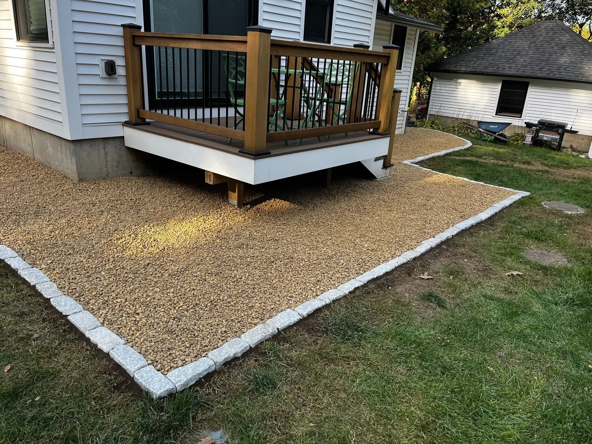 Tan gravel patio with granite edging bordering wooden deck with black metal railings