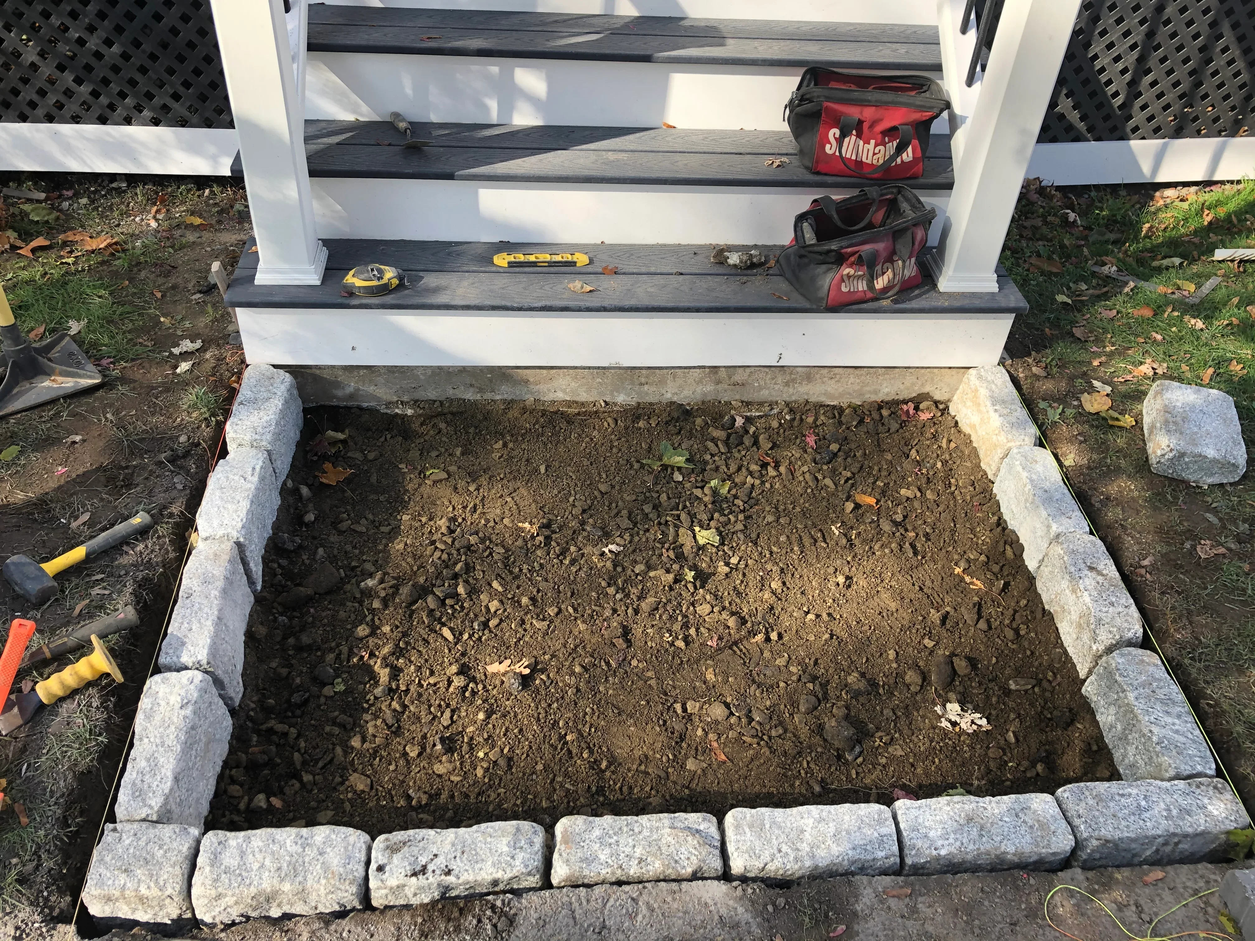 Raised garden bed with gray granite stone edging and compacted soil, adjacent to white deck with black railings
