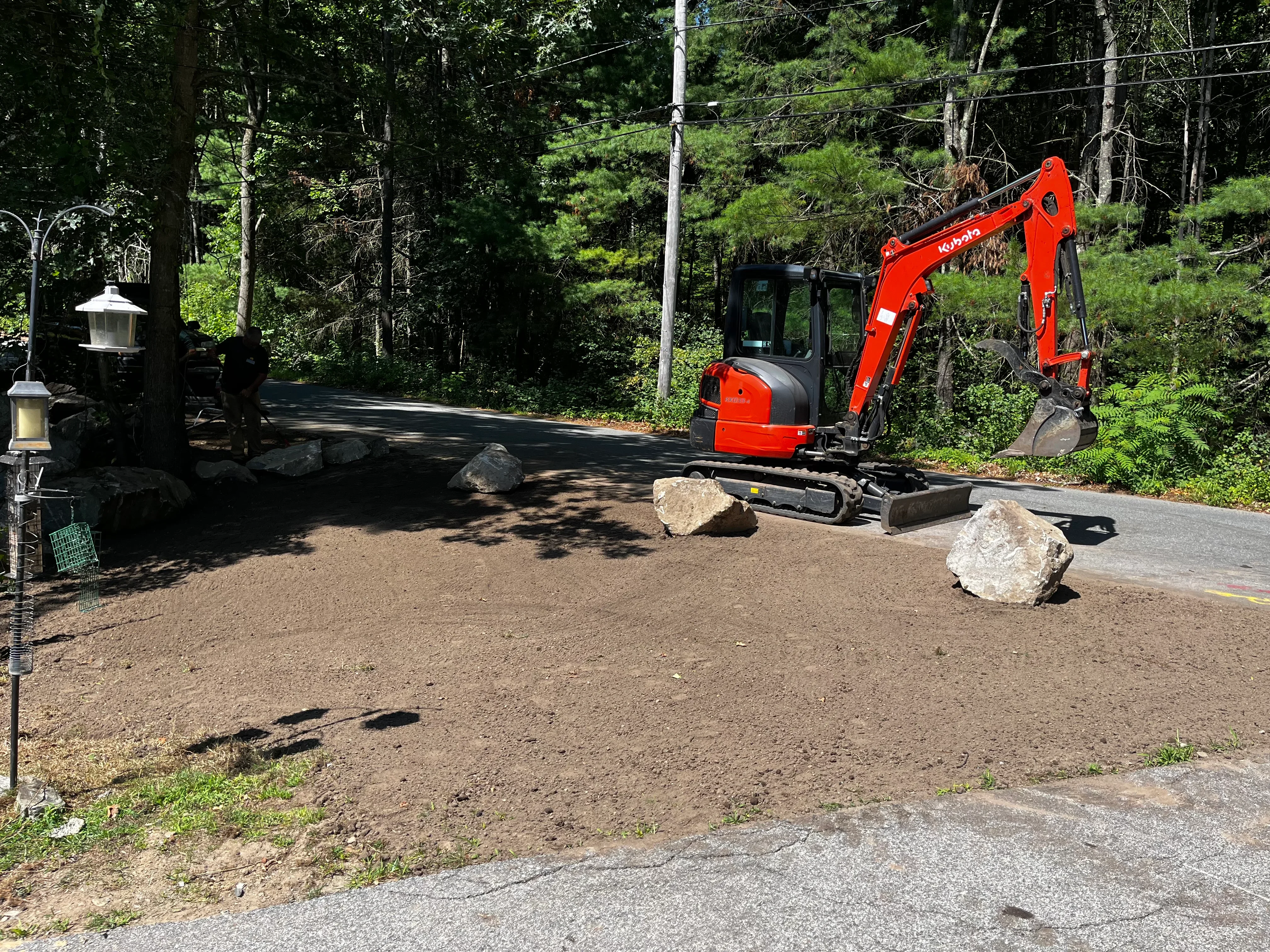 Red compact excavator grading driveway with decorative boulders and wooded backdrop
