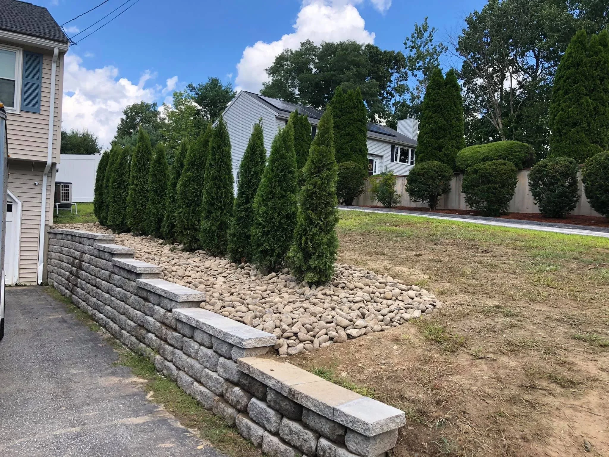 Stacked stone retaining wall with drainage rock and tall evergreen shrubs for privacy screening