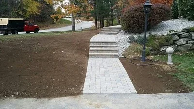 Newly installed gray paver walkway through prepared landscape bed with stone steps and lamp post