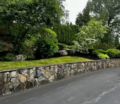 Stacked stone retaining wall along driveway with mature trees and landscaping above